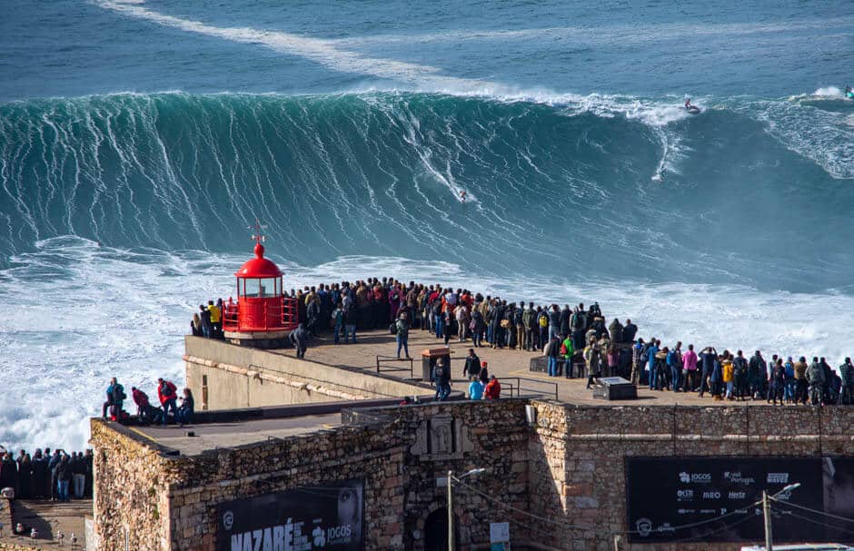 A giant wave approaches people standing on a platform at a lighthouse