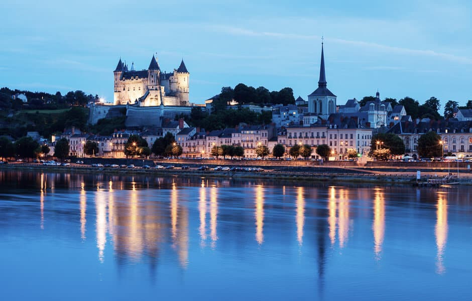 A castle with spires overlooking a town and river at blue hour