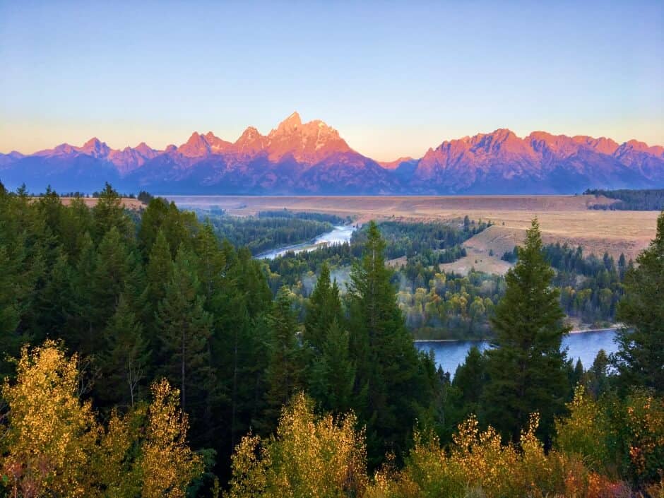 Mountains glowing in the sun act as a backdrop for a river and fall foliage in Grand Teton National Park, the best fall vacation destination in the U.S.