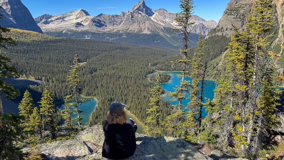 A woman sits on a mountain peak looking at lakes below with more mountains in the background