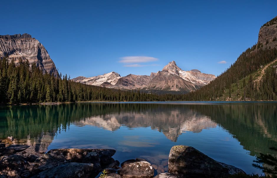A mountain with a pointy peak reflecting in a very blue lake surrounded by alpine fir trees.