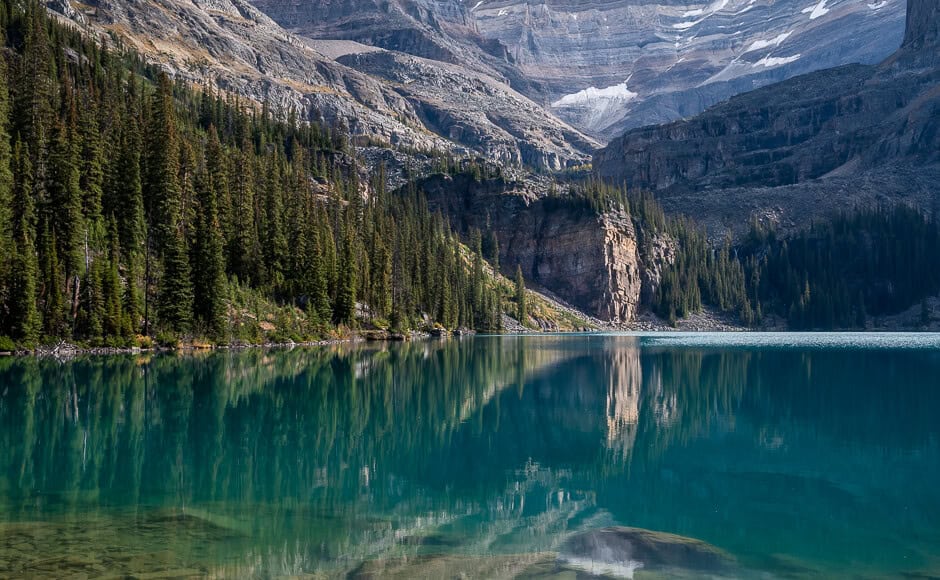 A mountain scene with the sun shining on a rock face reflecting on a lake with a glacier in the background in Yoho National Park