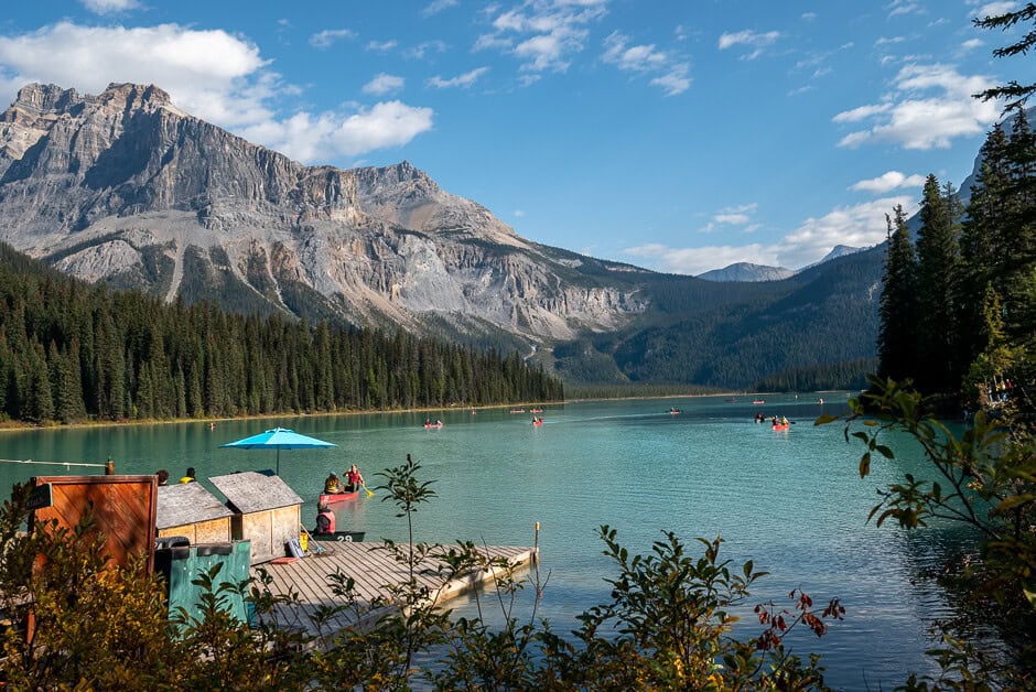 A dock on a blue lake with canoes in the water and a mountain, blue sky backdrop.