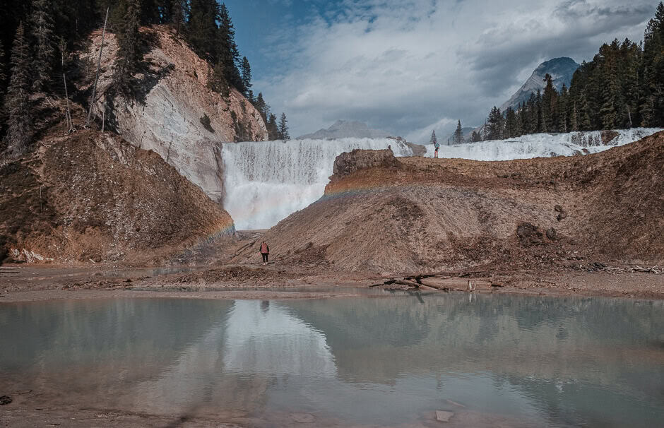 Am extremely wide waterfall in the mountains with a large mound of dirt in front and one person standing near it.